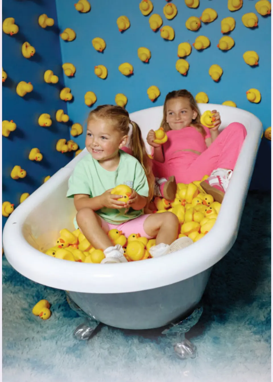 Two girls pose in a clawfoot tub surrounded by rubber ducks.